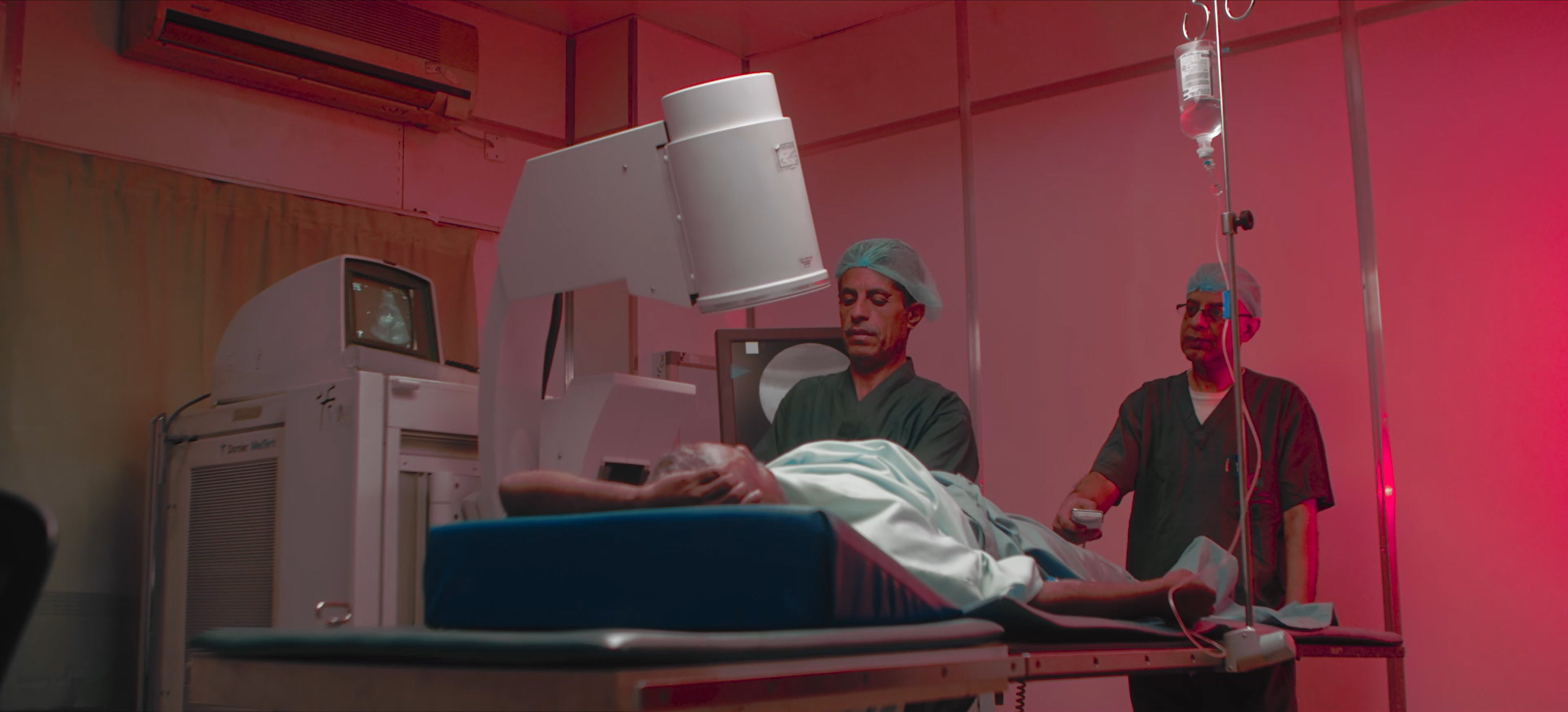 Two clinicians attending to a patient beside medical imaging equipment in a red-lit room