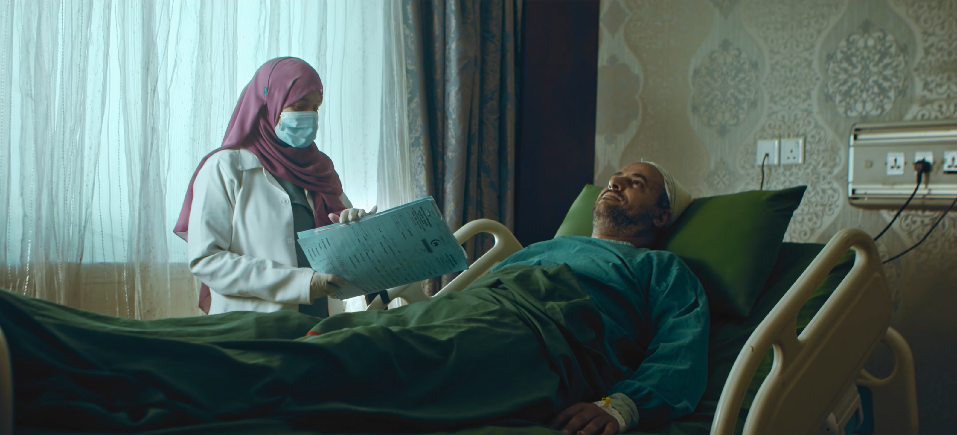 A masked clinician with a patient chart beside a man resting in a hospital bed by the window